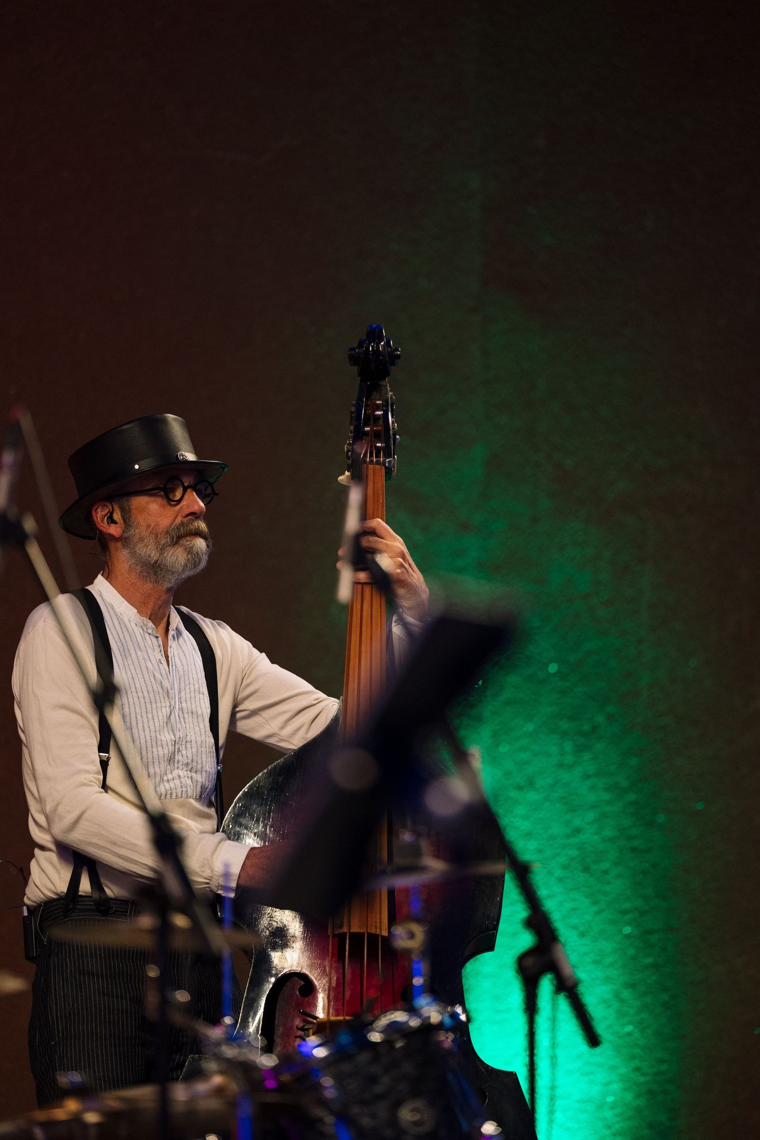 Bearded jazz musician in hat and suspenders plays upright bass on dimly lit stage with green lighting.