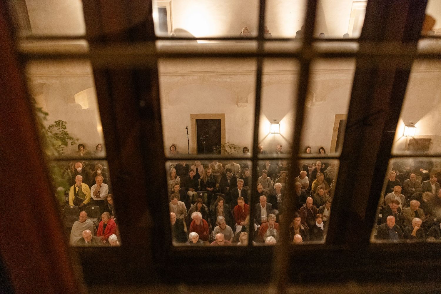 Crowd of people gathered in a warmly lit historic hall, viewed through a barred window from outside.