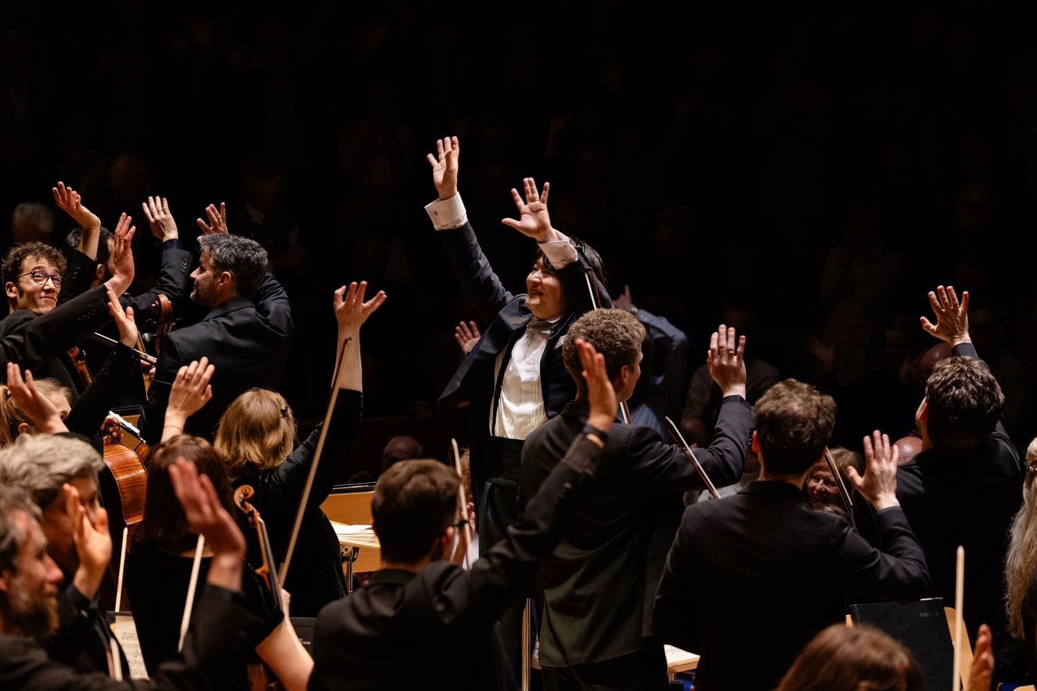 Orchestra conductor and musicians raise hands during a dramatic classical music performance on a concert hall stage.