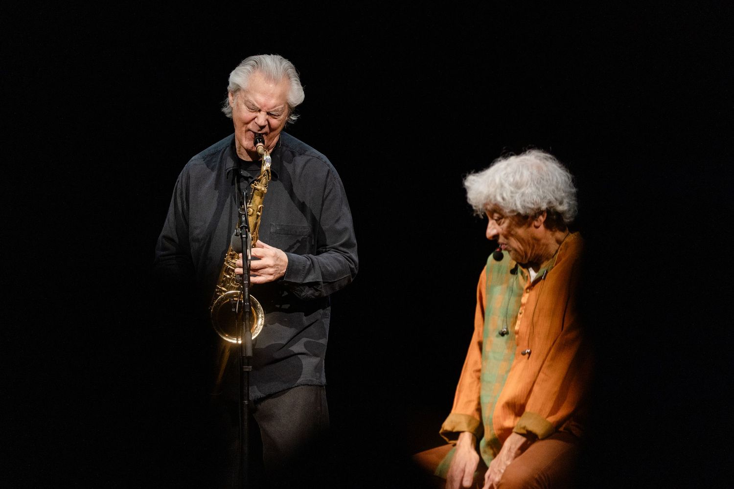 Two musicians perform on stage, one playing saxophone in black attire, the other seated in an orange outfit.