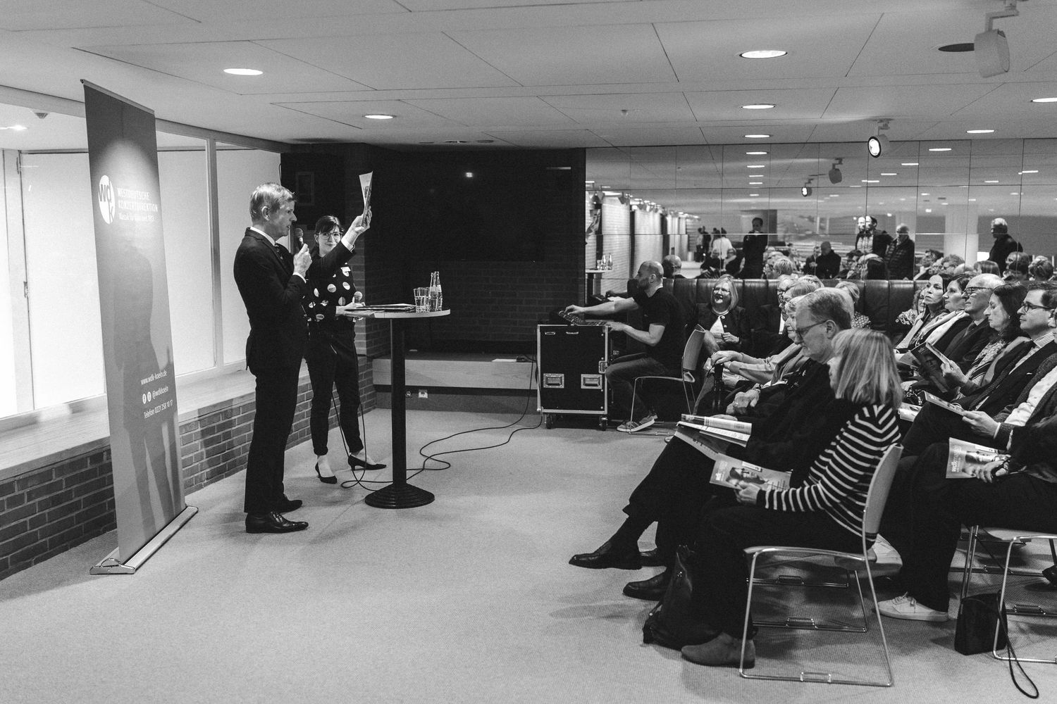 Two presenters speak at a podium before an engaged seated audience in a modern conference room setting.