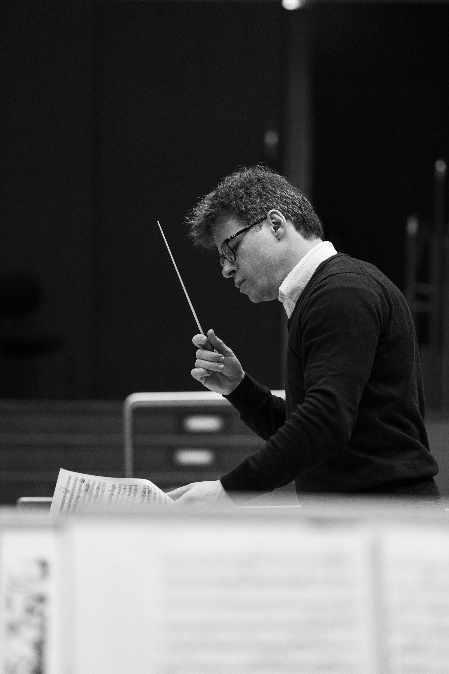 Black and white photo of a male orchestra conductor holding a baton while reading sheet music during rehearsal.