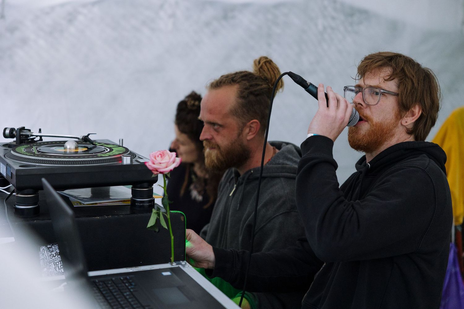 Two men DJing at an outdoor event, one drinking from a bottle, with a turntable and laptop on the table.