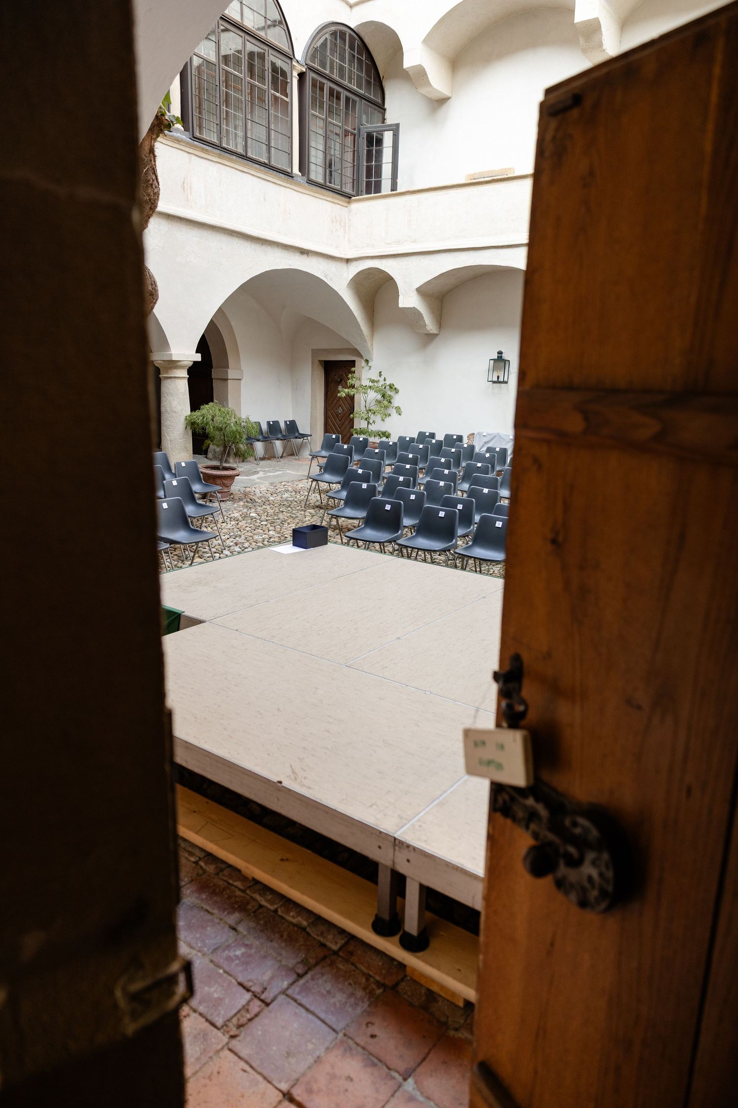 Courtyard venue with blue chairs arranged around a stage, viewed through a wooden door in a white arched building.