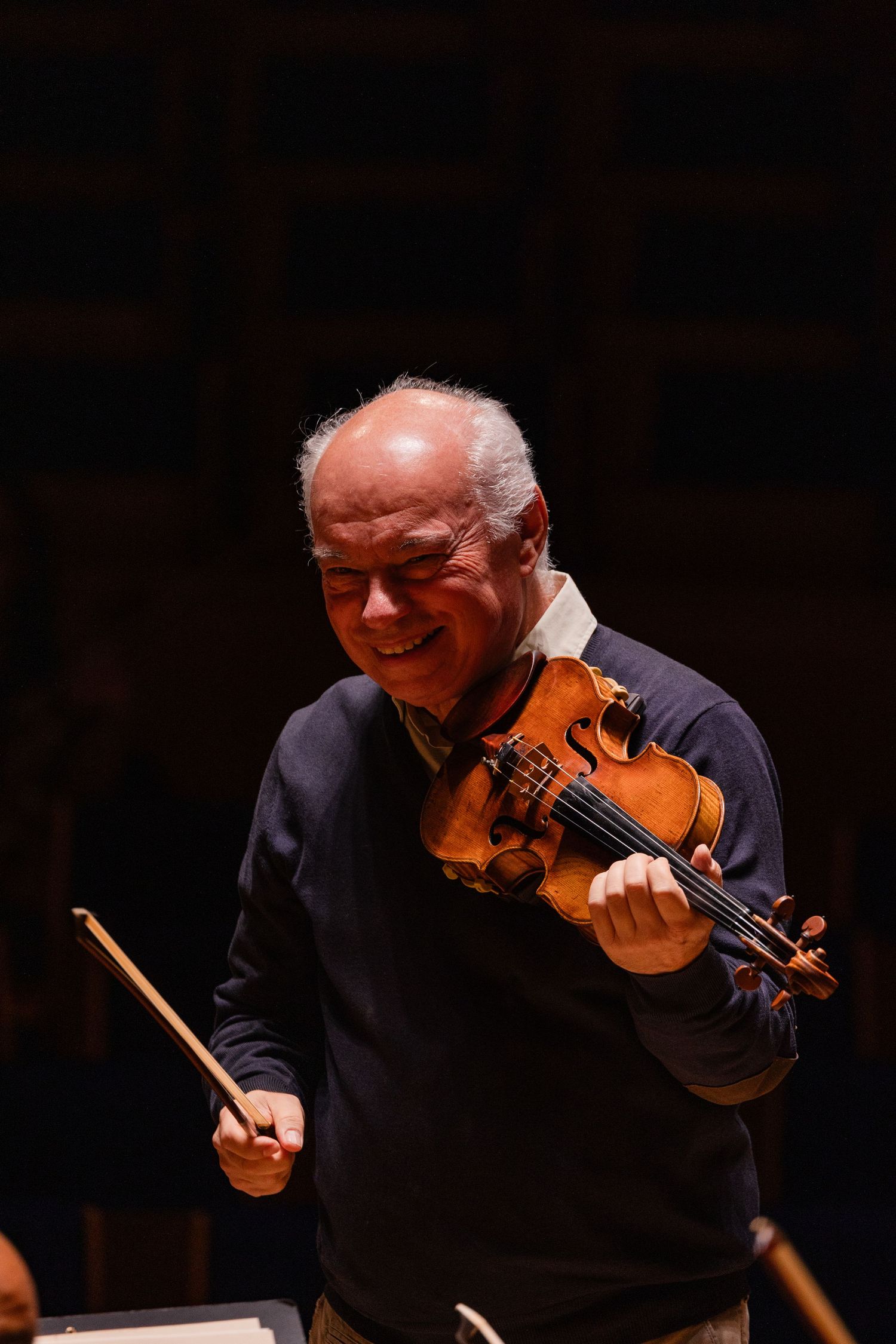 Elderly violinist smiling while playing violin and holding bow on dark concert stage.