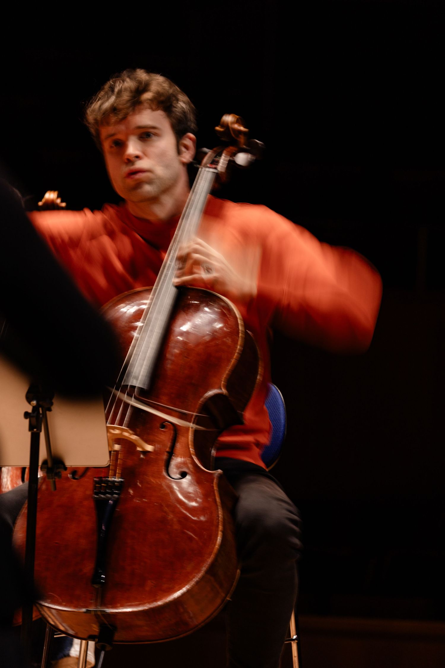 Young male cellist in red shirt performs passionately on stage, motion blur capturing his expressive playing technique.