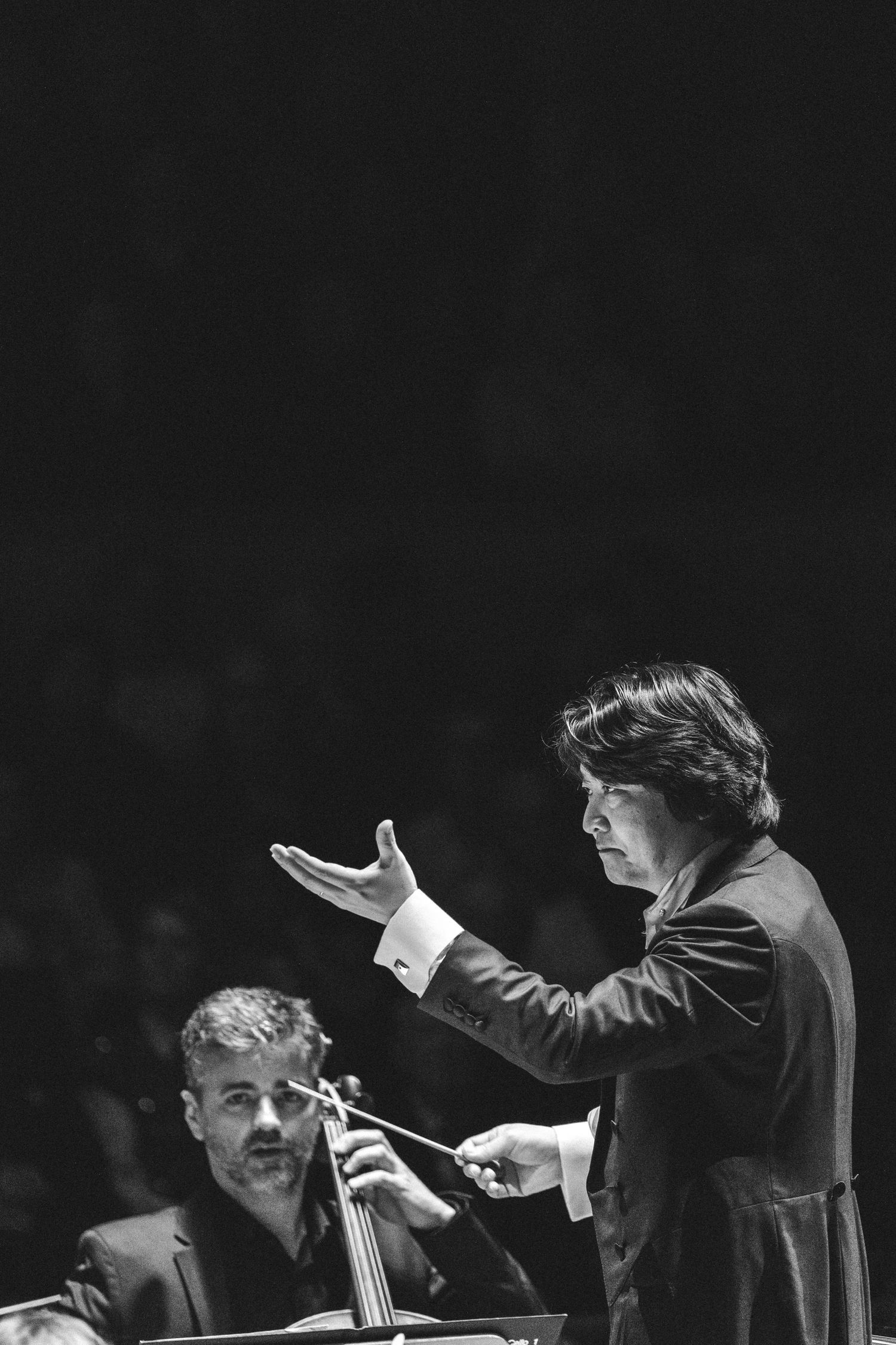 Black and white photo of an orchestra conductor gesturing with baton while musician plays cello in background.