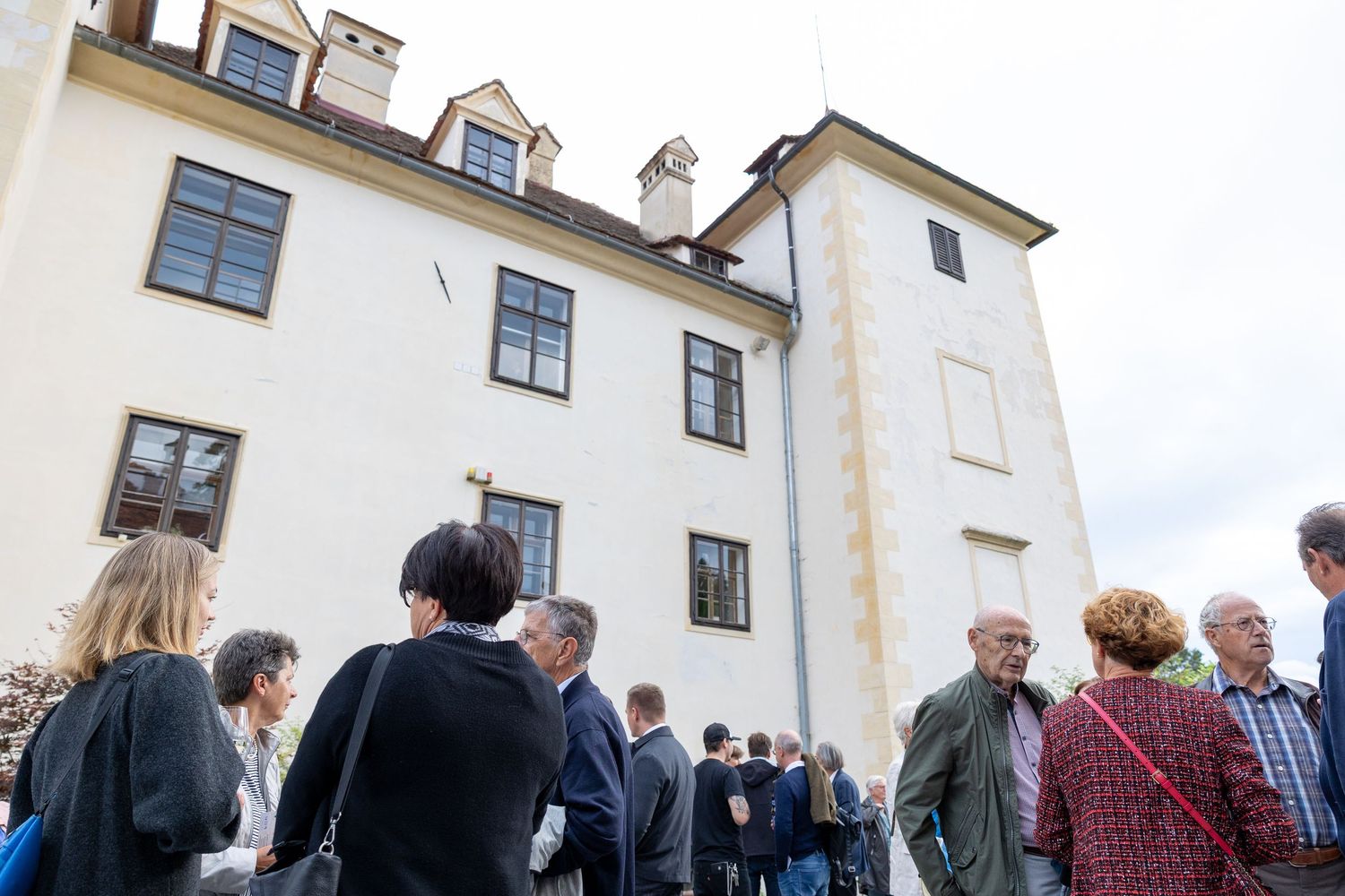 Guests gather outside a historic white European manor building during an outdoor social event on a cloudy day.