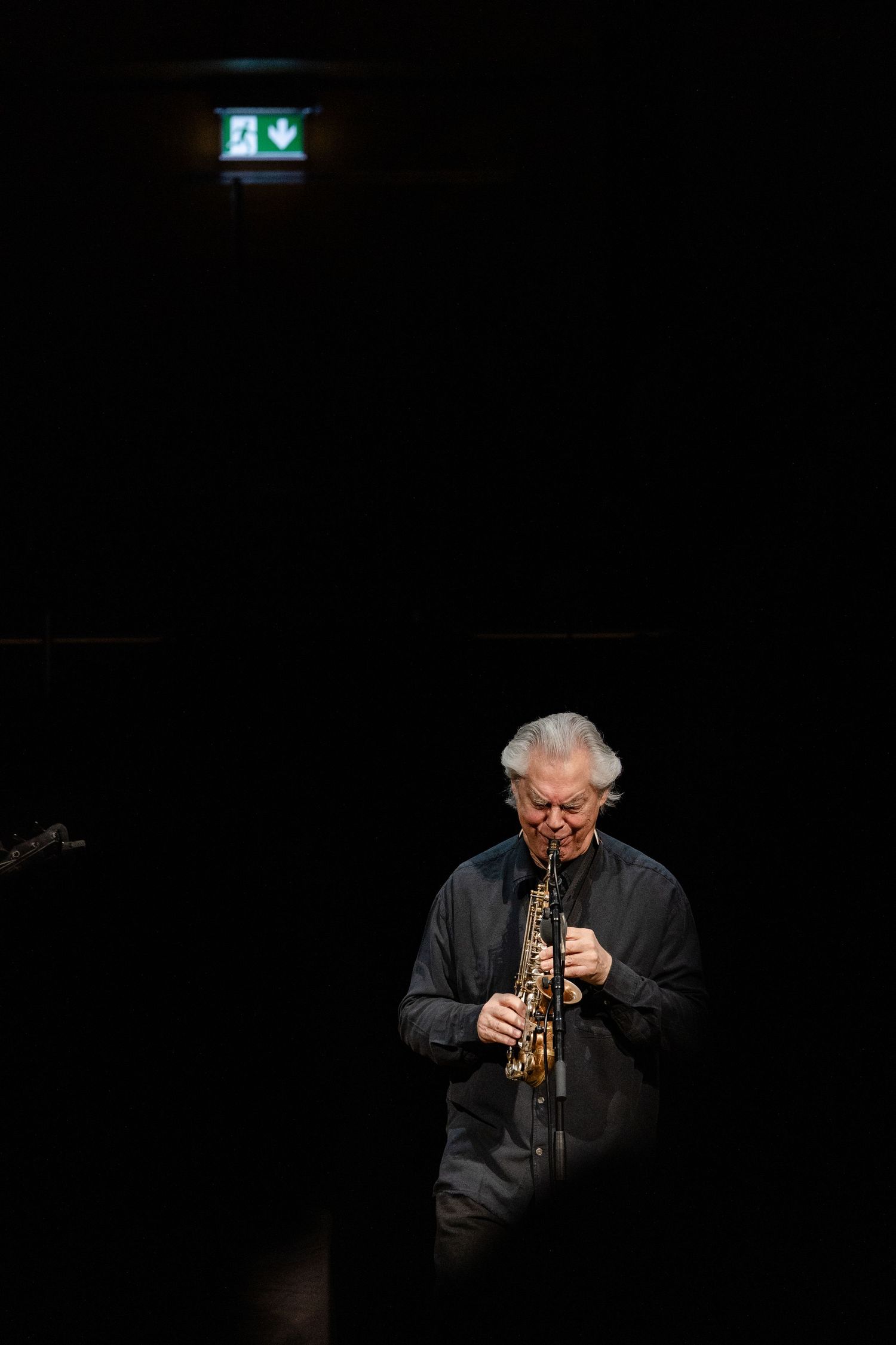 Musician in dark jacket performs on stage playing a brass instrument against a black background.
