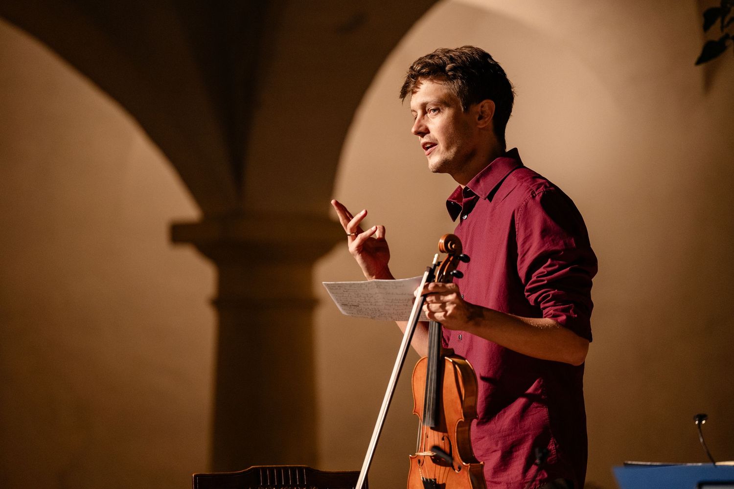 Young male violinist holding violin and bow while speaking at a music performance in an arched venue.