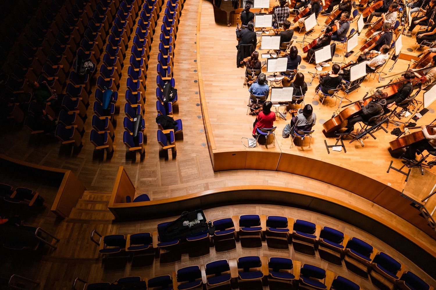 Aerial view of an orchestra performing on stage in a concert hall with curved rows of blue seats.