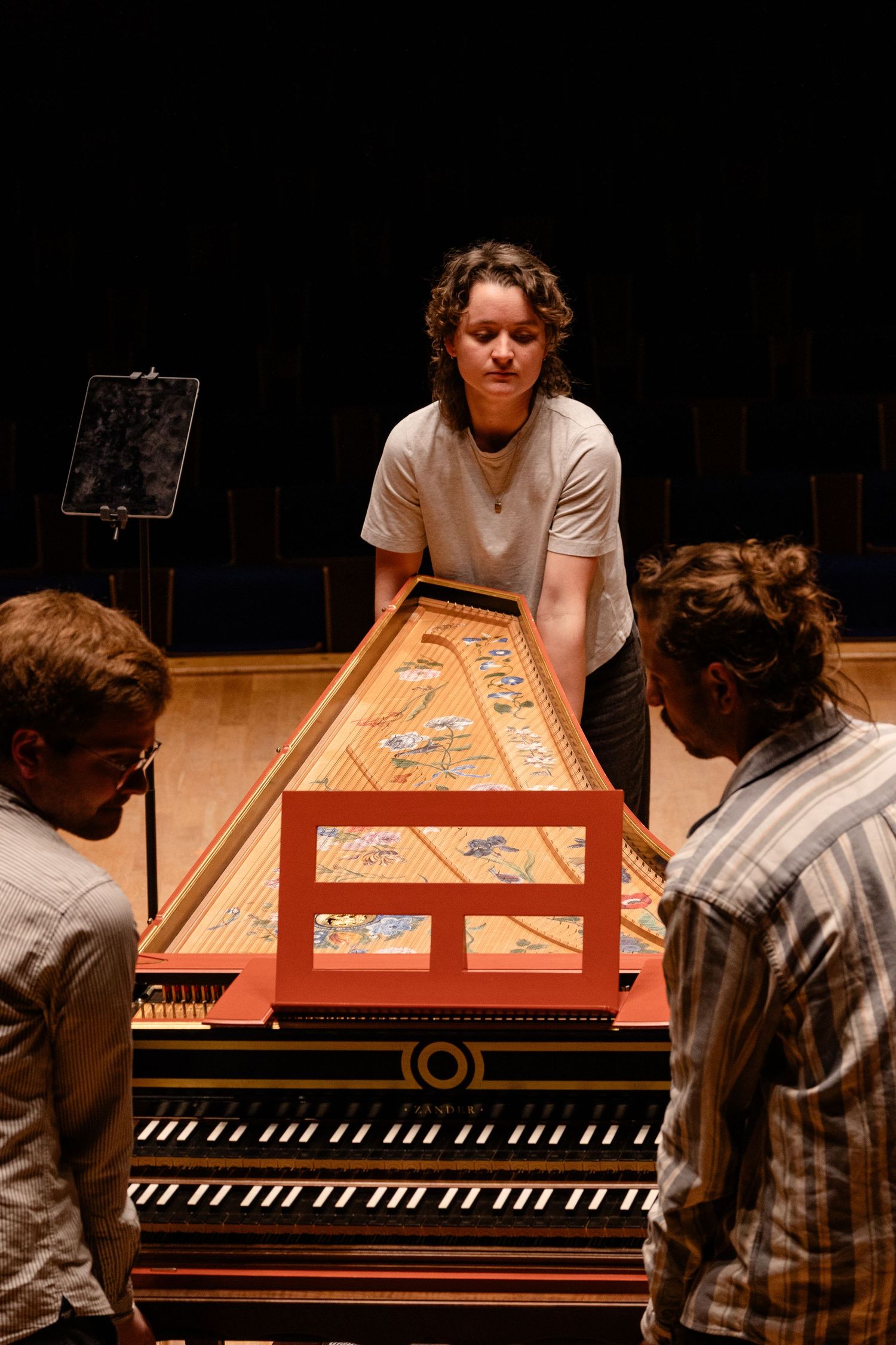 Three musicians examine an ornate harpsichord on a concert stage, with decorative painted lid open.