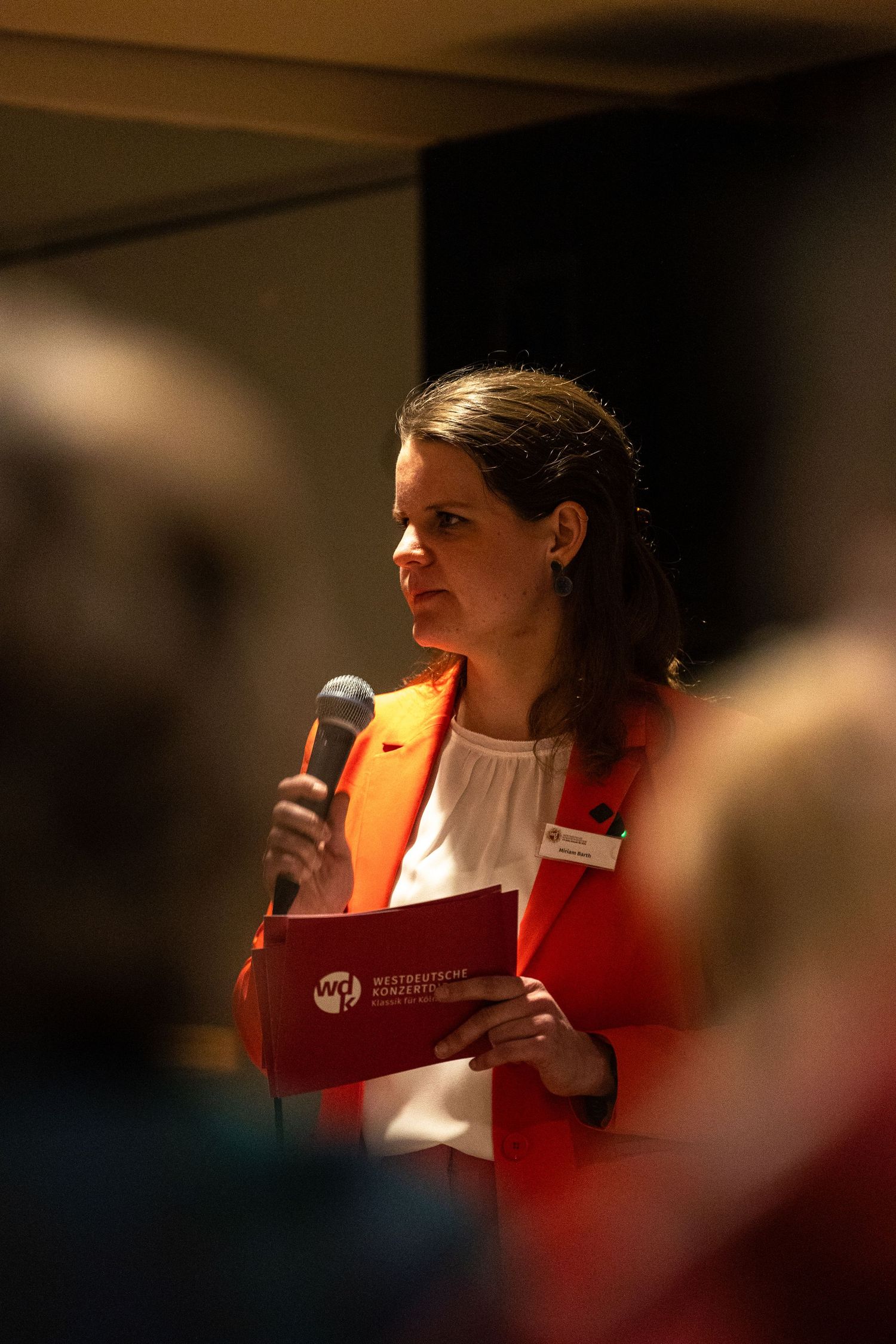 Woman in red blazer speaking at event, holding microphone and red folder with Westdeutsche Konzerte logo.