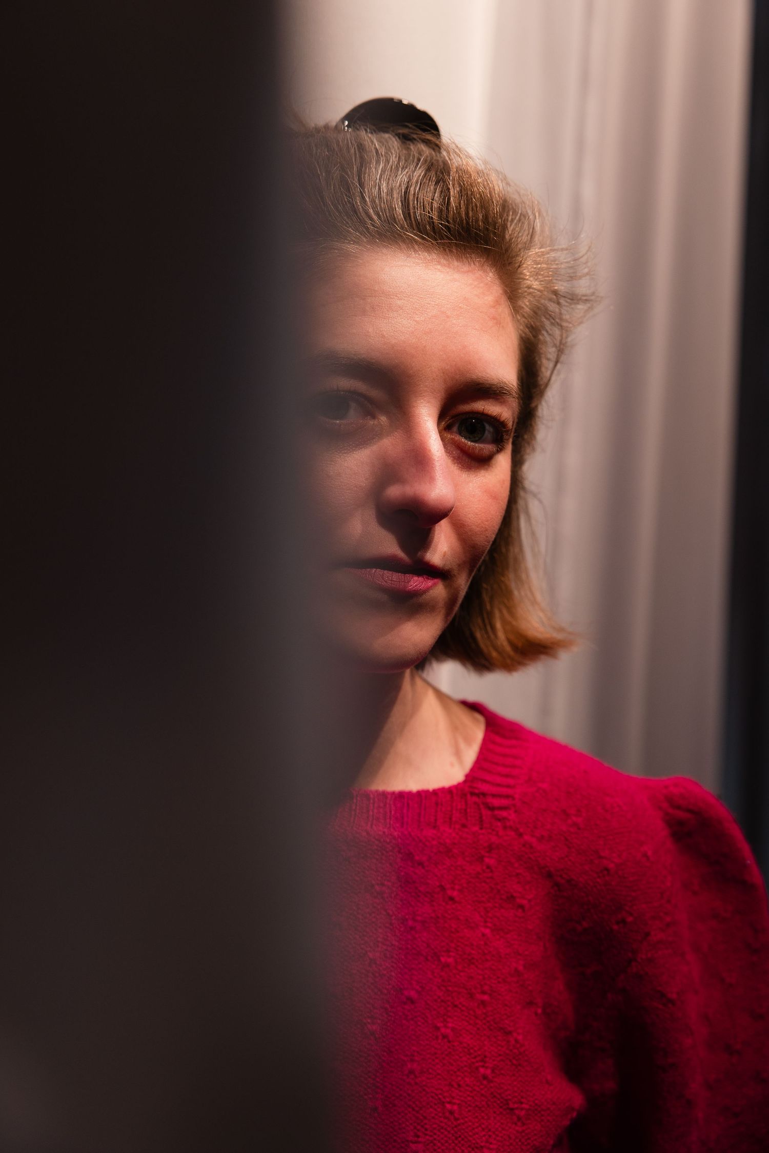 A woman in a red sweater with short blonde hair gazes thoughtfully past a dark foreground element near a curtained window.