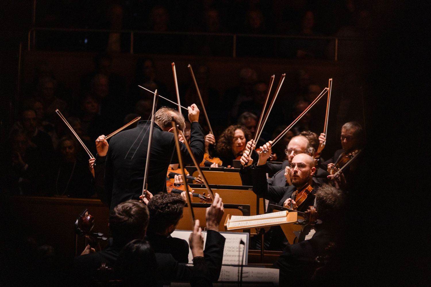 Orchestra performing in a dimly lit concert hall with string section visible.