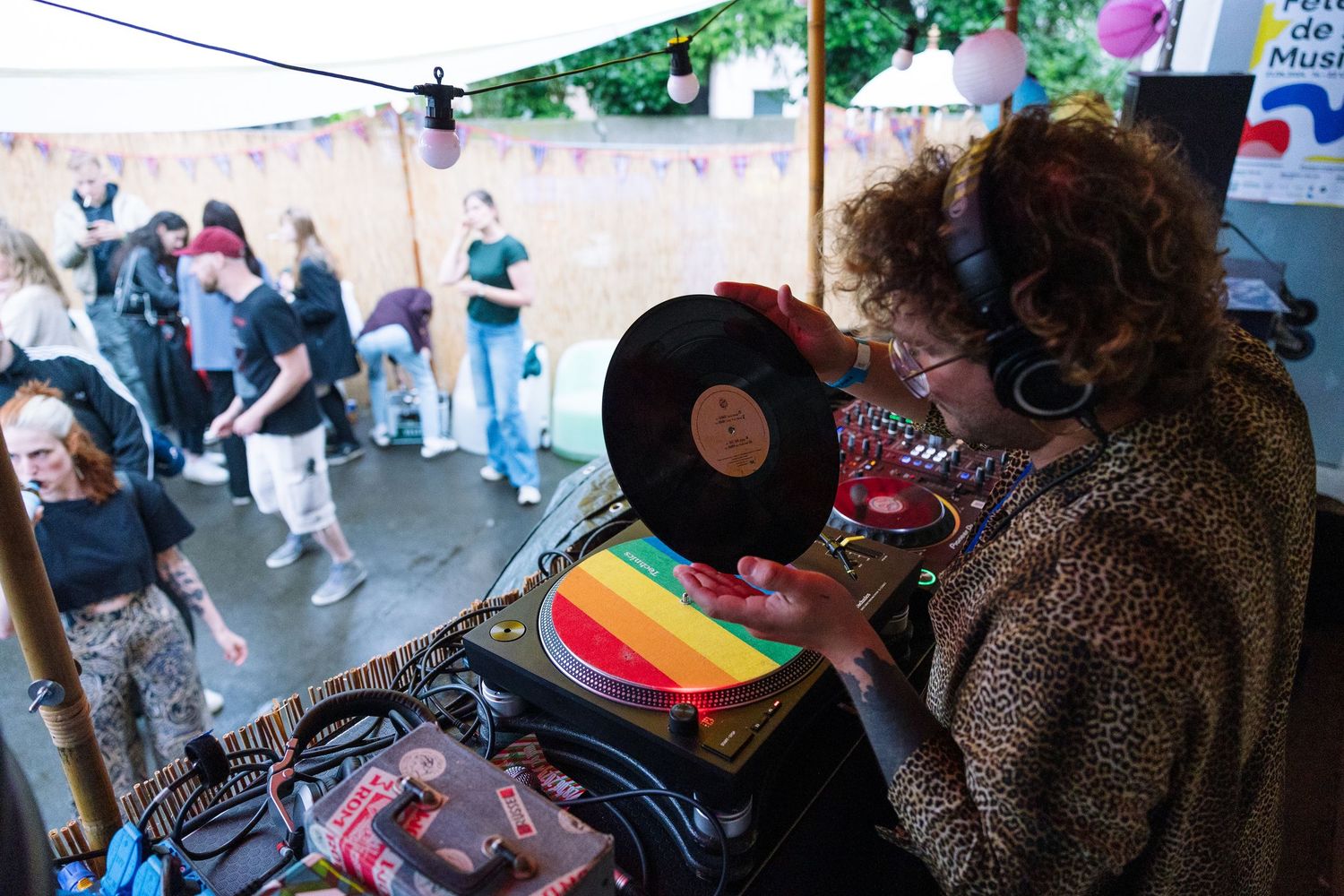 A DJ with curly hair and headphones holds a vinyl record at an outdoor music festival with dancers in background.