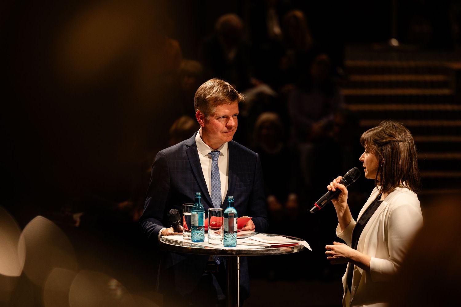 A man in a navy suit and a woman in white speak at a podium during a formal panel discussion event.