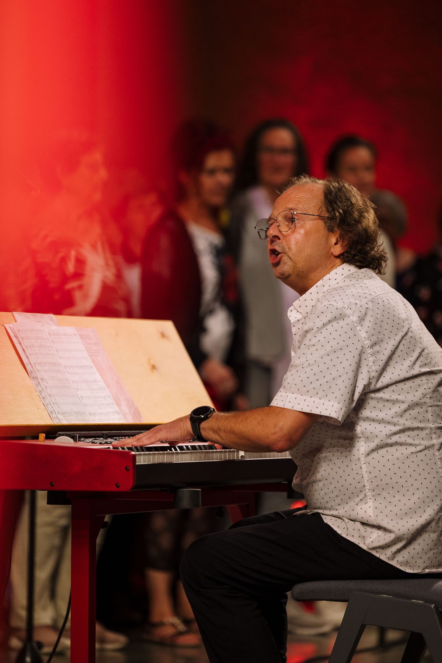 Man playing red keyboard piano on stage, singing from sheet music, with audience in background under red lighting.