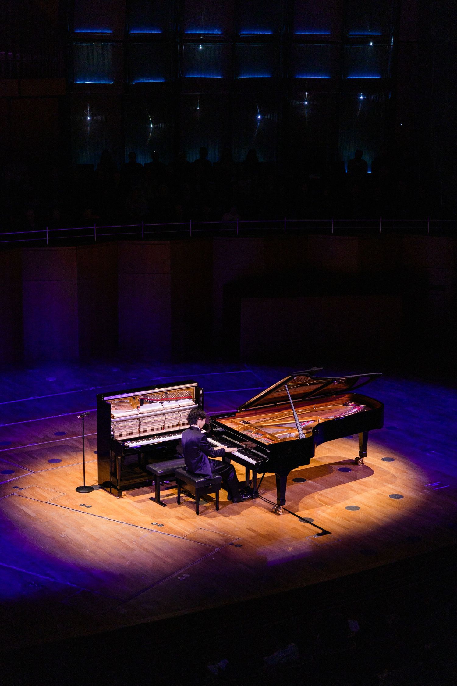 Pianist performing solo on a grand piano under warm spotlight on a concert hall stage with blue ambient lighting.