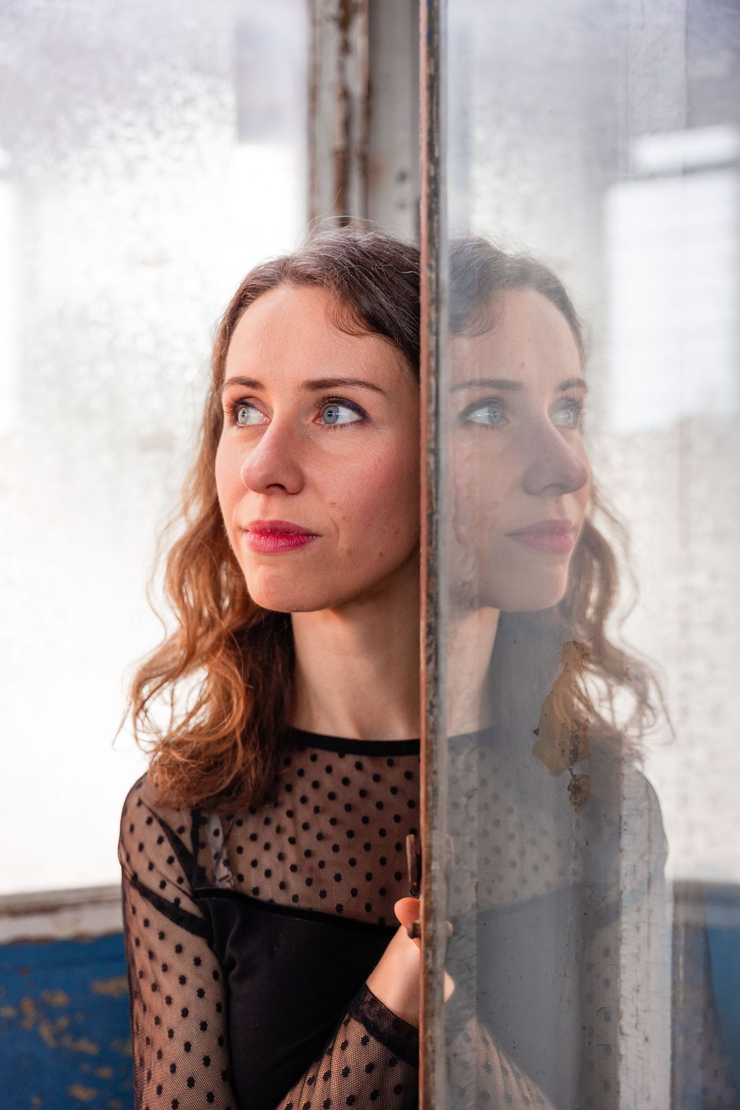 A young woman with curly brown hair in a black polka dot top gazes upward beside a mirror reflecting her profile.