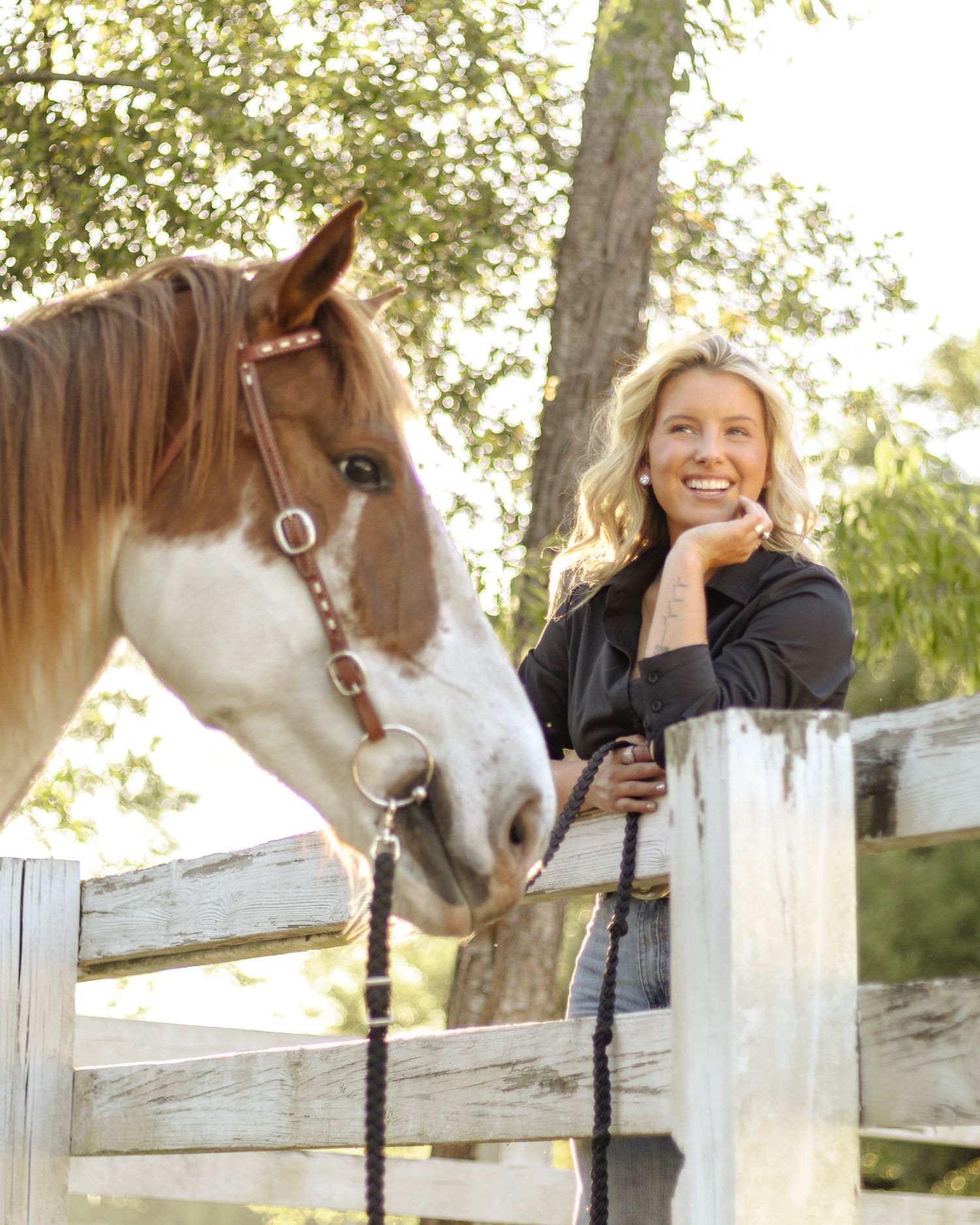 Smiling blonde woman in a black shirt leans on a white wooden fence beside a brown and white horse wearing a leather halter.