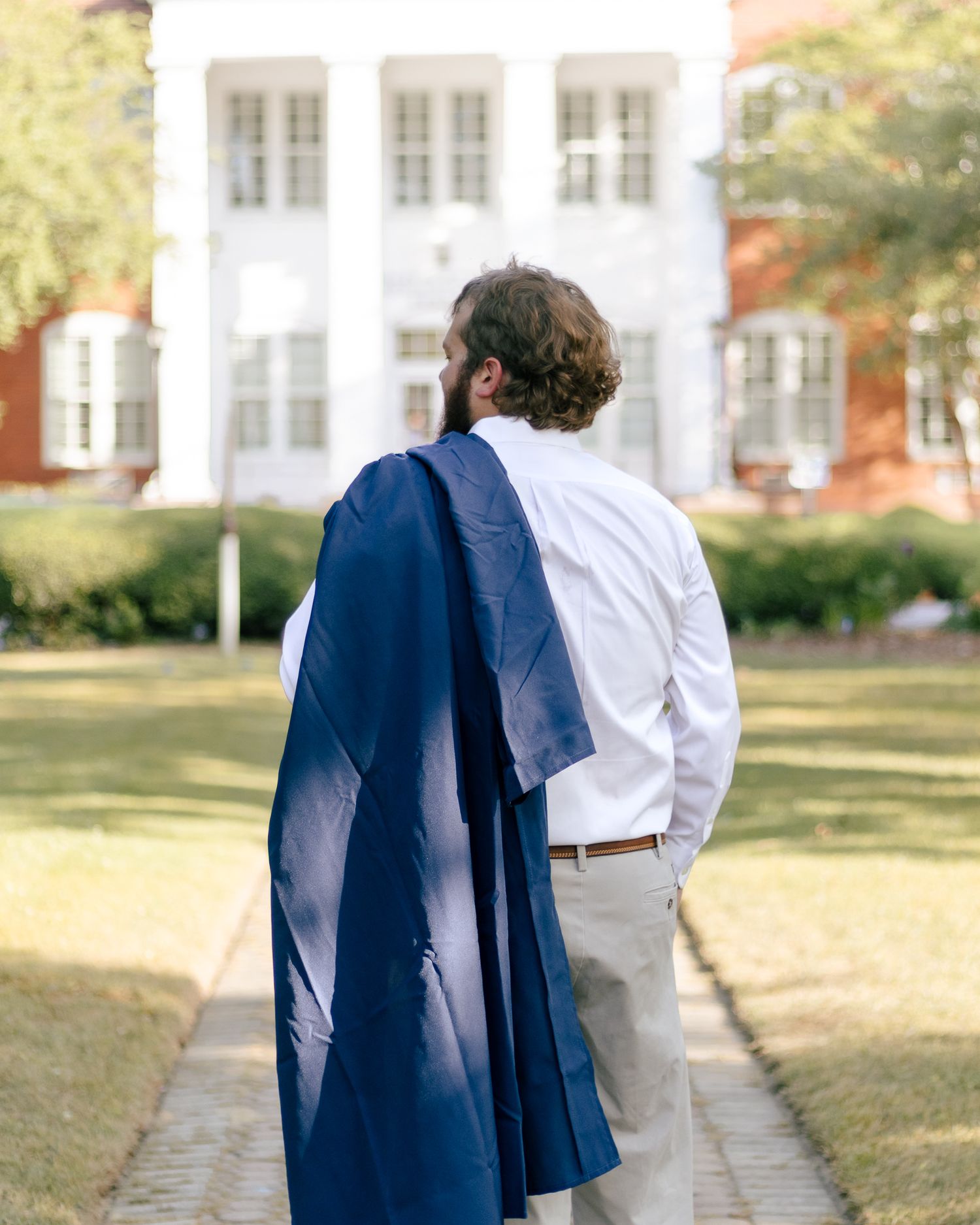 Graduate in a white shirt and khakis walks a brick path toward white-columned campus buildings, a navy gown draped over his shoulder.