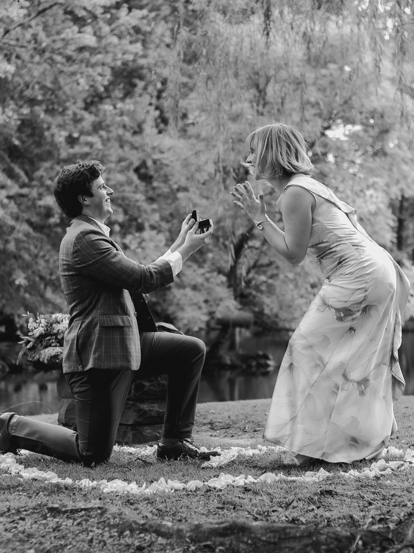 Black and white photo of a man kneeling with an open ring box as a woman in a floral dress reacts with joy at a park proposal.