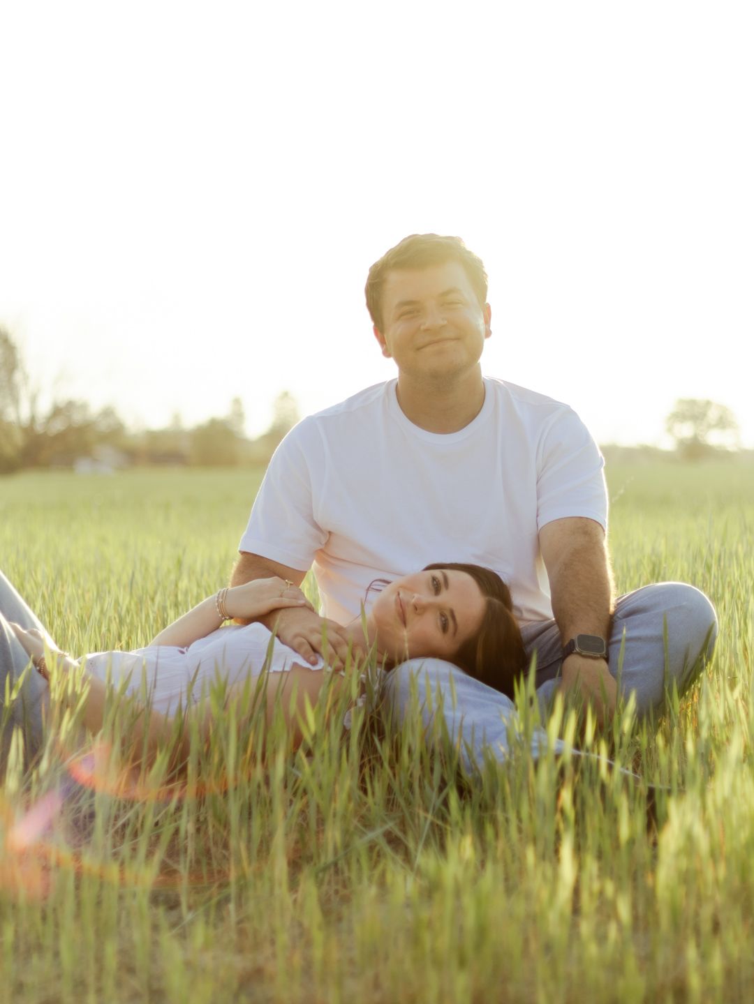 Smiling man in a white t-shirt sits in a green grass field at golden hour while a woman rests her head on his lap.