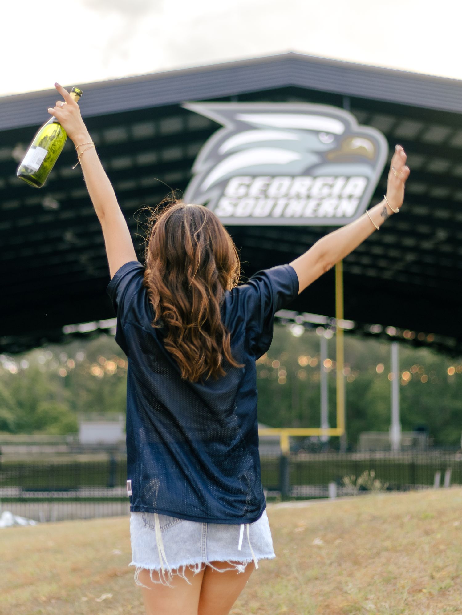 Woman in a navy jersey and denim shorts raises her arms and a wine bottle outside a Georgia Southern stadium sign.