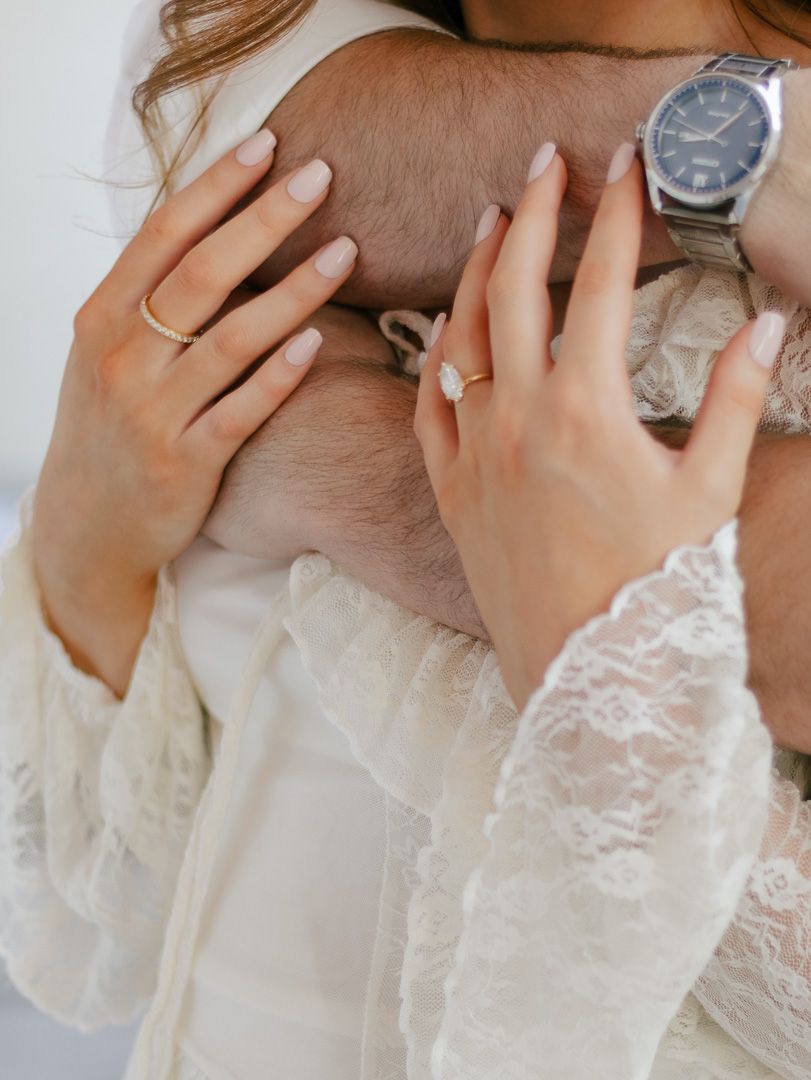 Woman in a cream lace-sleeved dress wearing a diamond engagement ring as she wraps her arms around her partner, who wears a wristwatch.