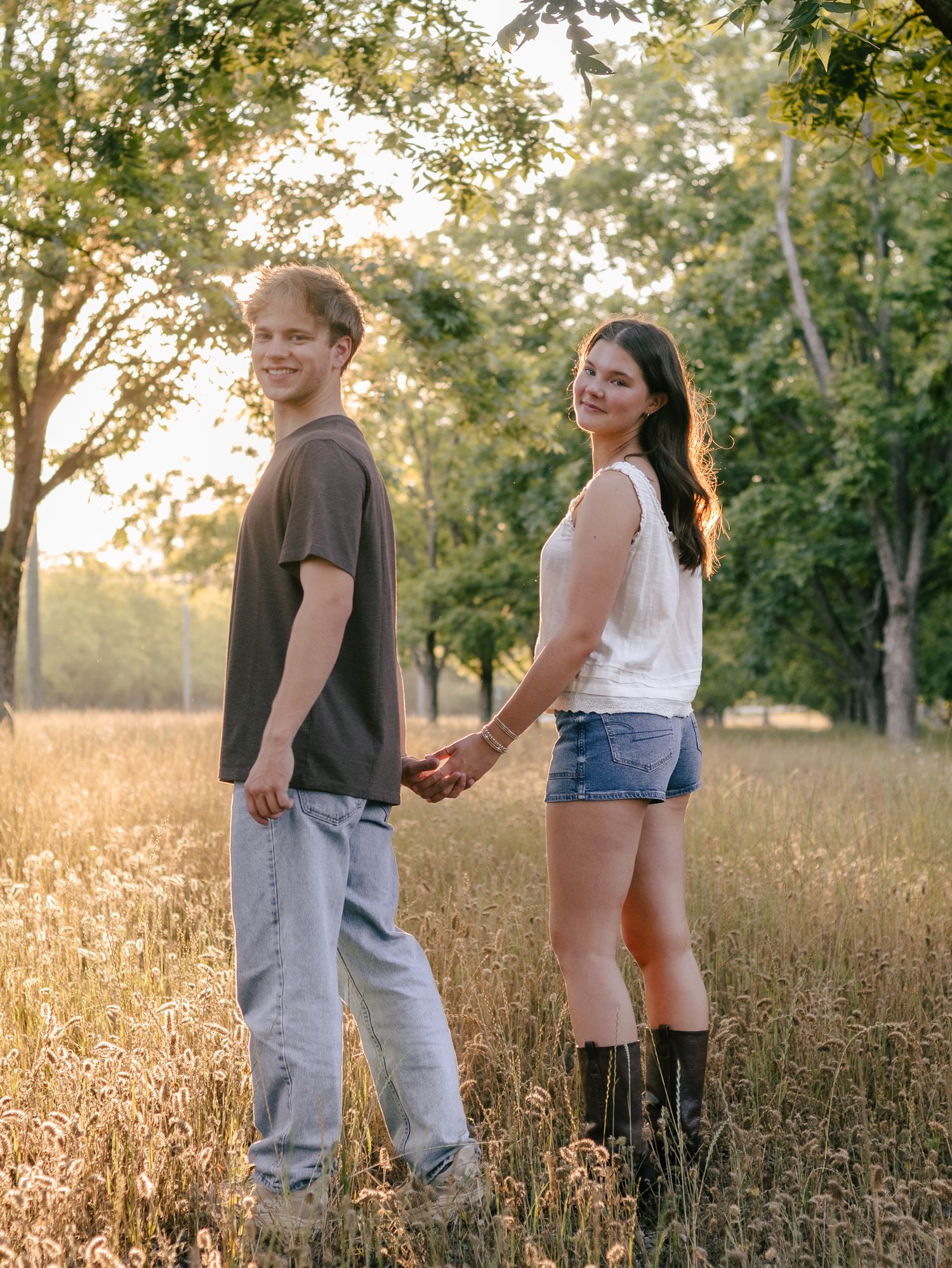 Young couple holding hands and smiling back at the camera in a golden grassy field with sunlit trees at sunset.