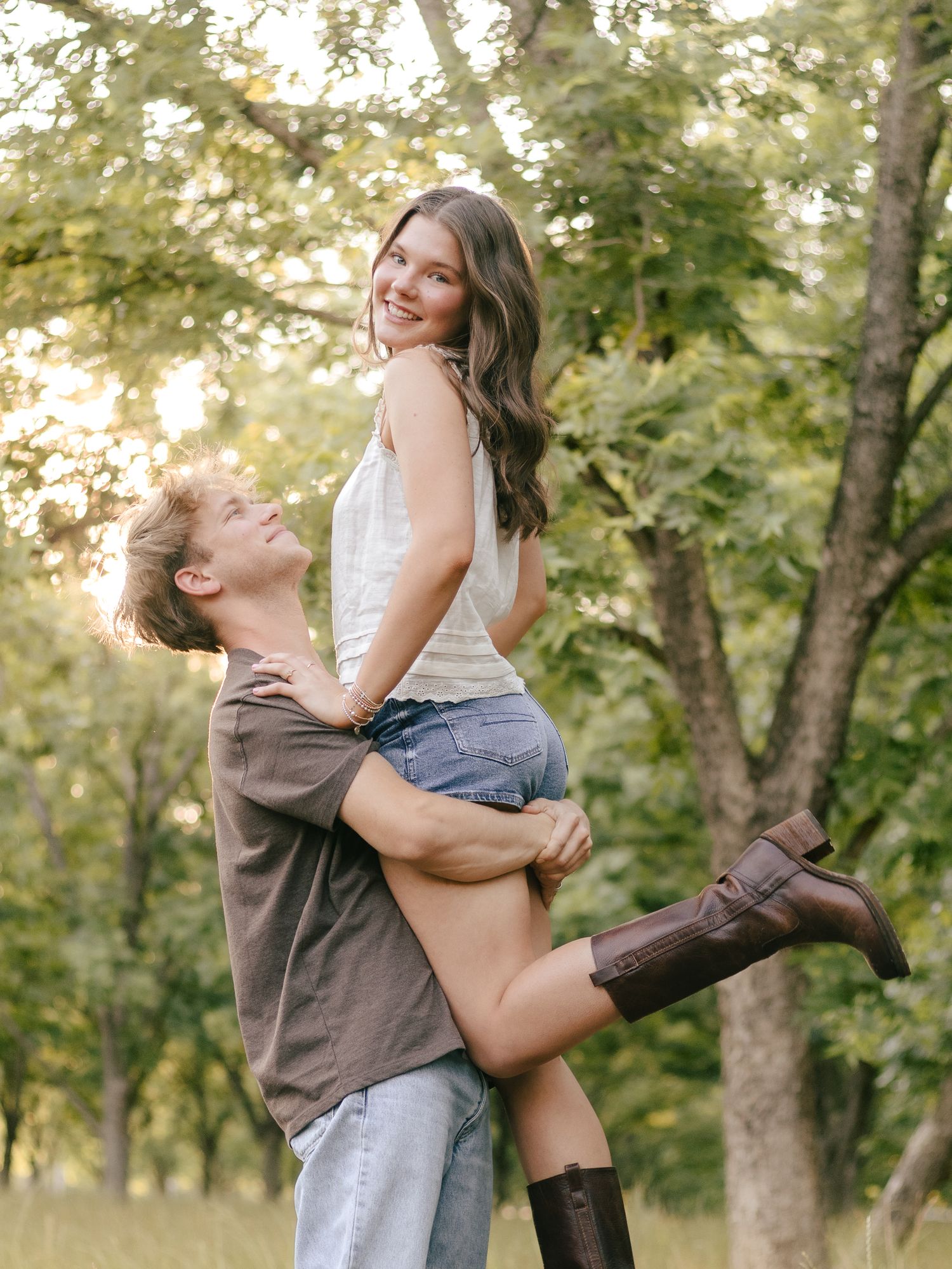 Smiling couple in an orchard as a man lifts a woman in a white top, denim shorts and brown cowboy boots beneath sunlit trees.