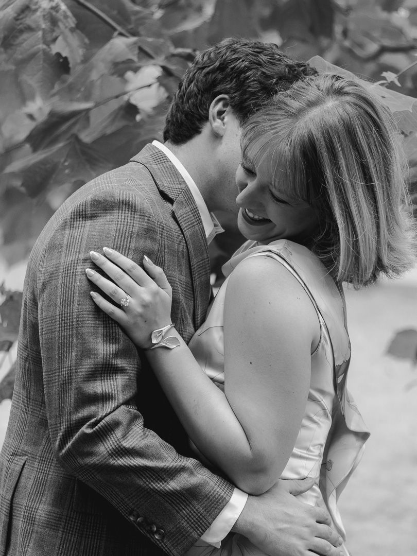 Black and white engagement photo of a smiling couple embracing closely as the man kisses her cheek beneath leafy trees.