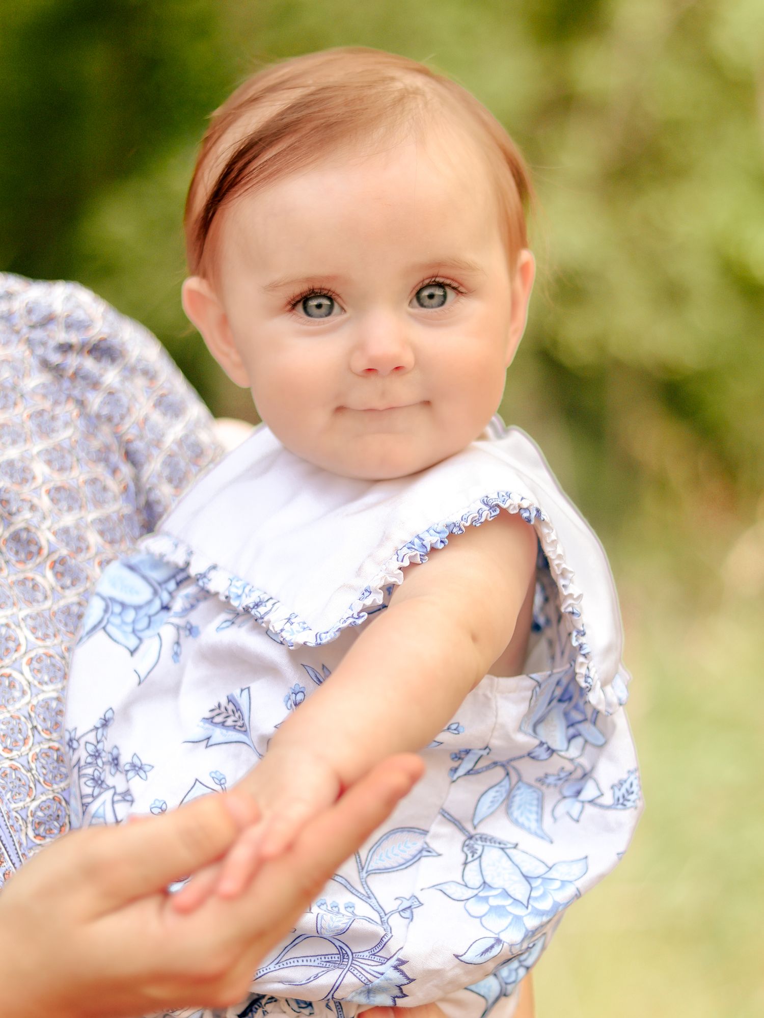 Smiling baby with light eyes wearing a white and blue floral ruffled outfit, held by an adult outdoors in a green park.