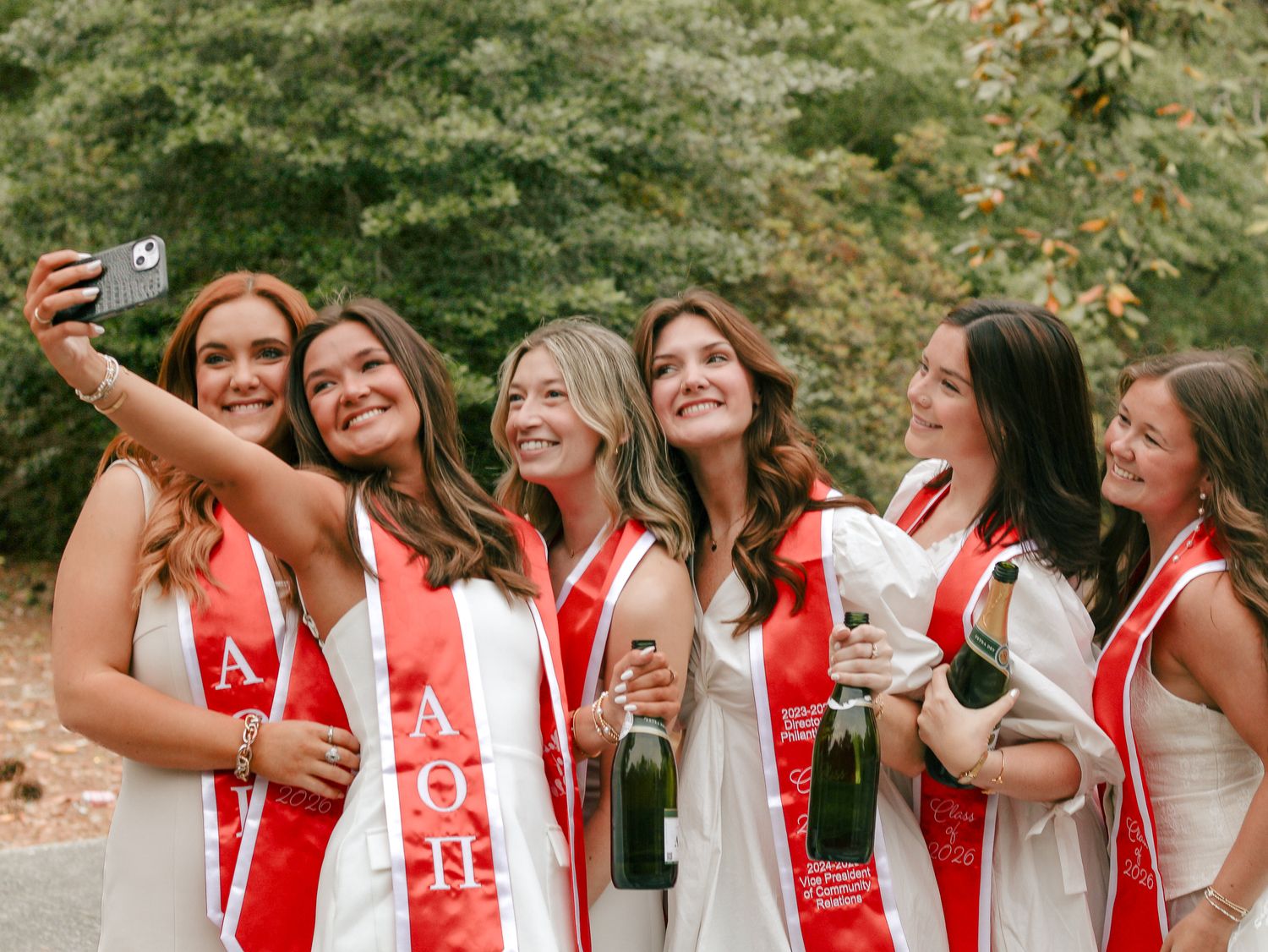 Group of smiling sorority graduates in white dresses and red sashes taking a selfie outdoors while holding champagne bottles.
