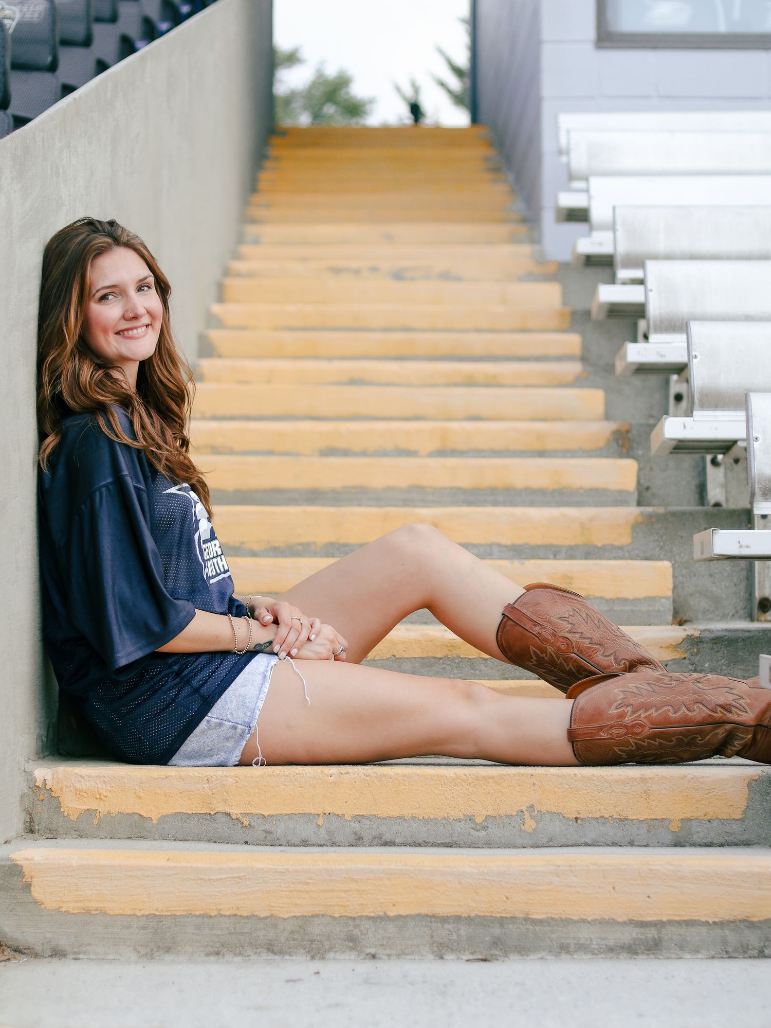 Smiling young woman with long brown hair sits on sunlit concrete stadium steps wearing a navy t-shirt, denim shorts and brown cowboy boots.