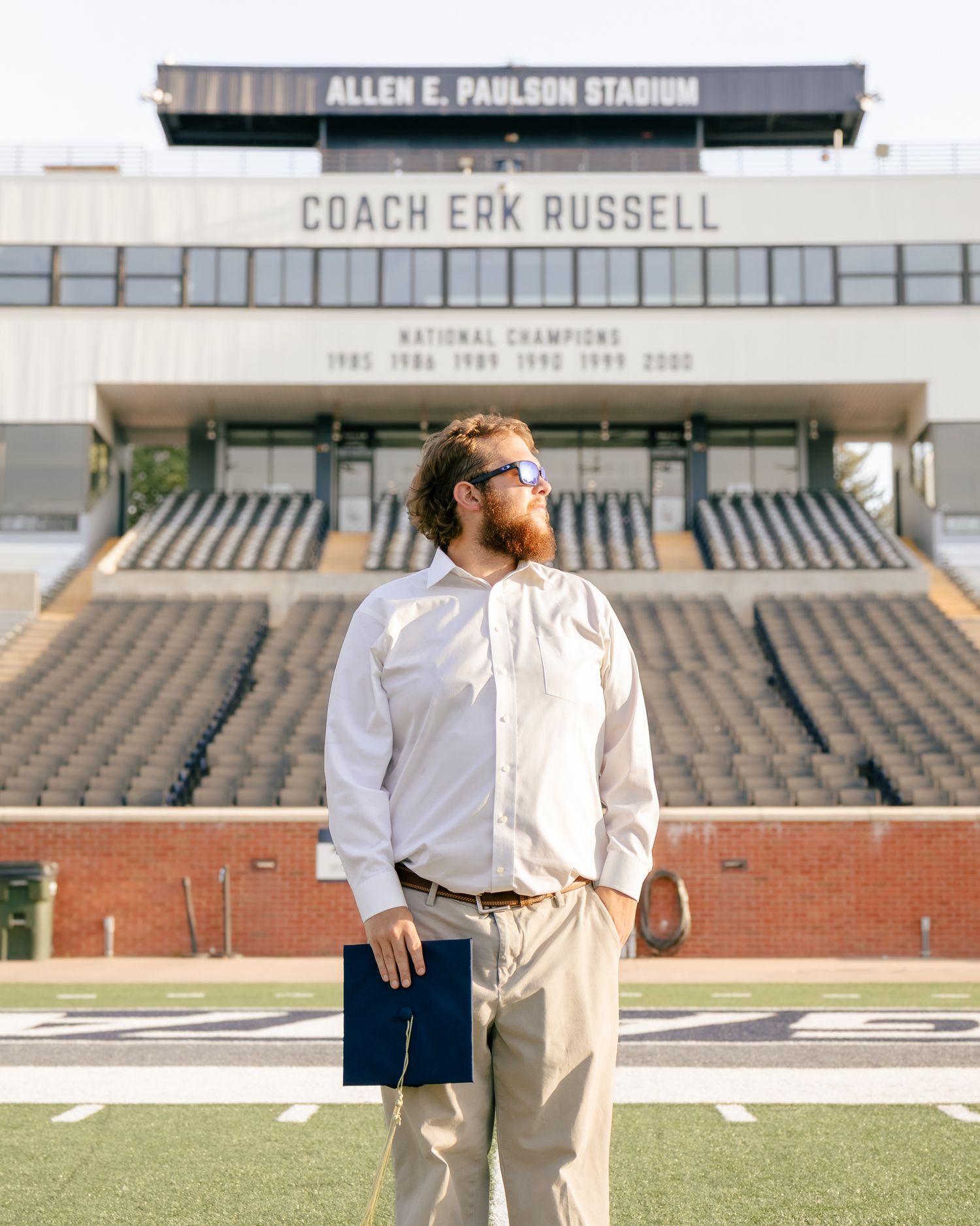 Bearded graduate in sunglasses, white dress shirt and khakis holds a diploma cover on the field at Allen E. Paulson Stadium.