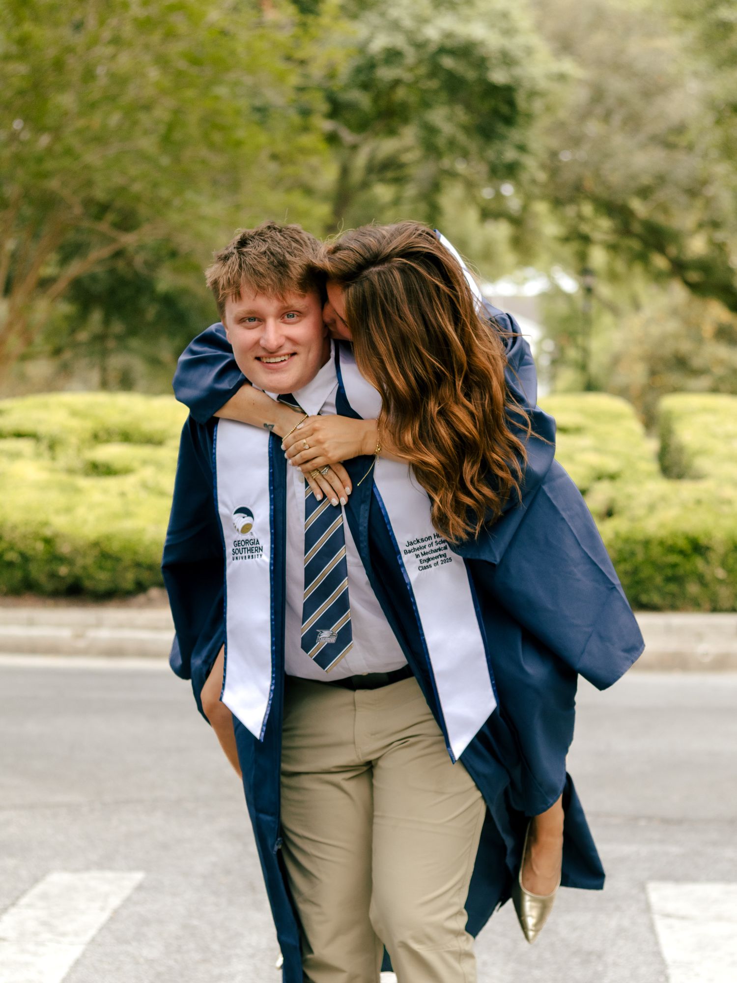 A smiling graduate in a blue gown and white stole is hugged from behind by a woman with long brown hair on a tree-lined street.