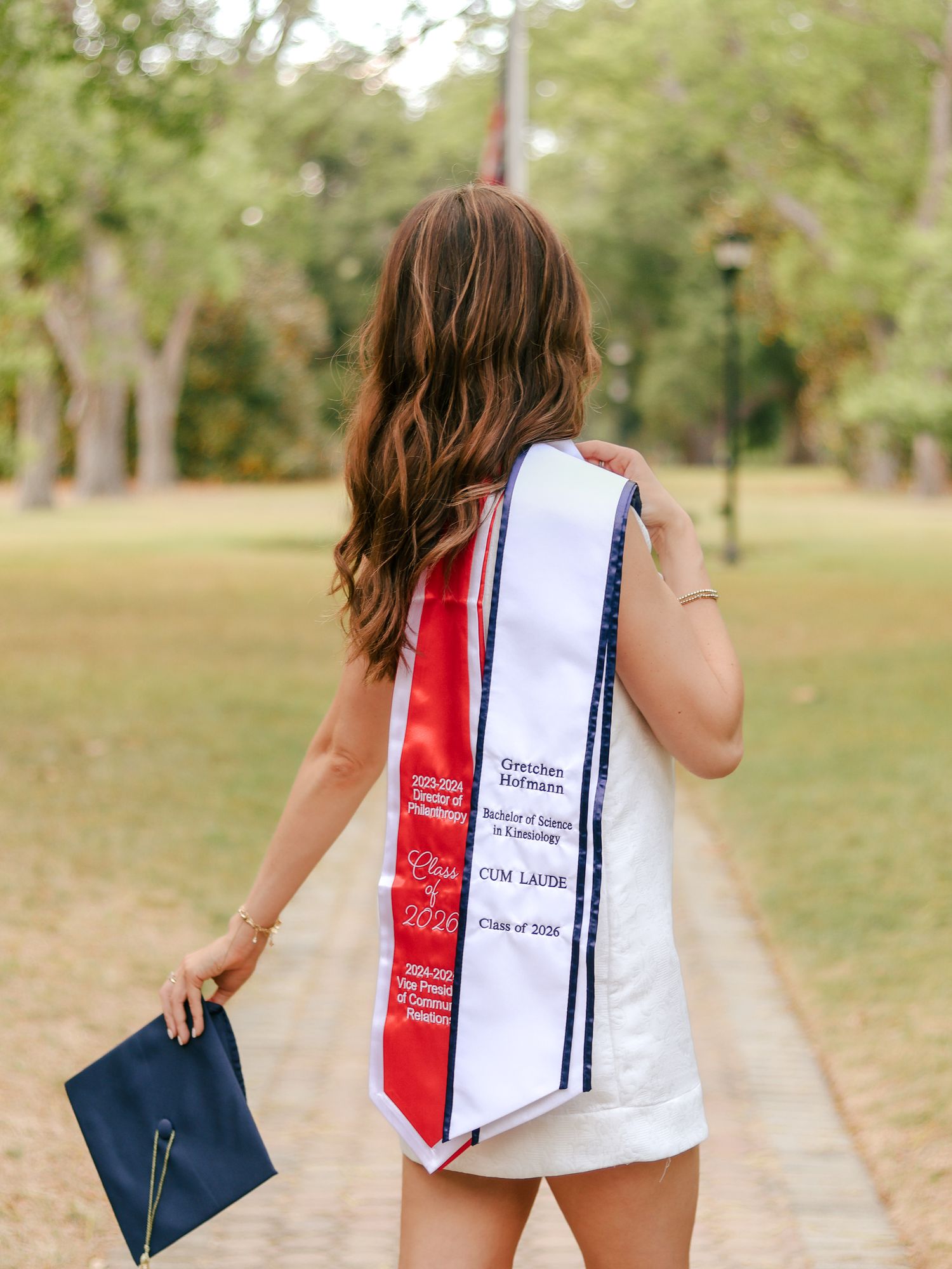 Graduate with long wavy hair faces away, wearing a white dress and a red and white cum laude graduation sash, holding a navy cap outdoors.