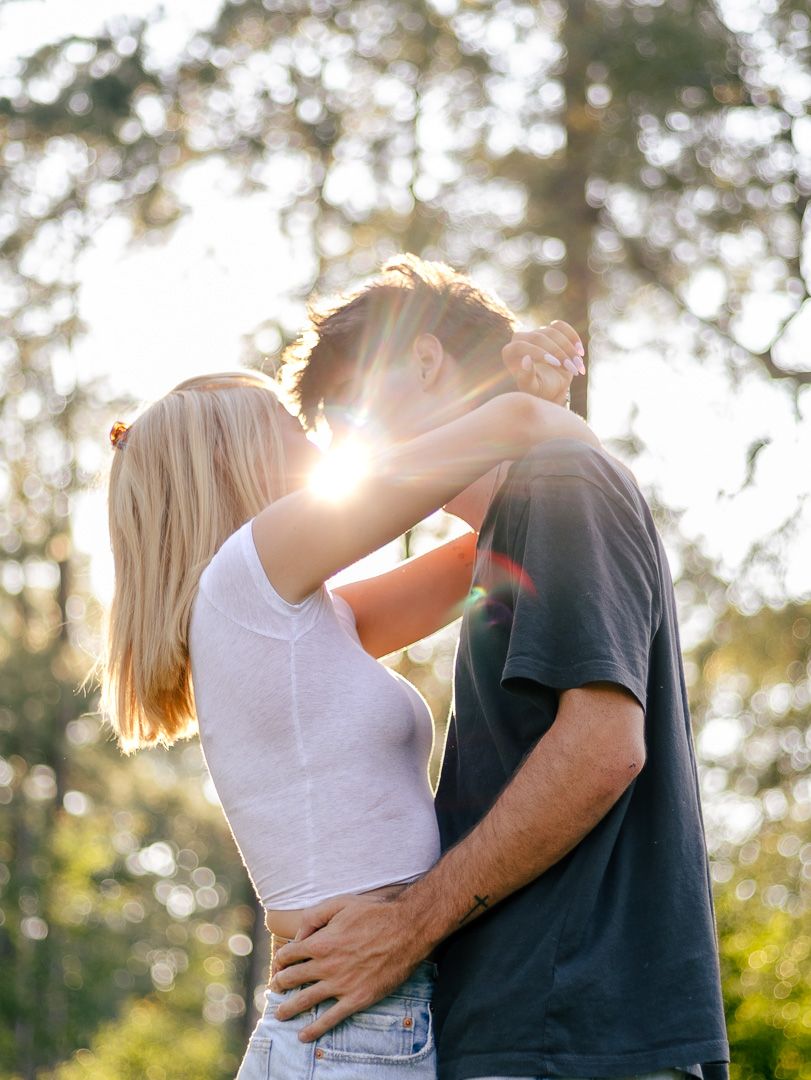 A blonde woman in a white top embraces a man in a dark t-shirt as golden sunlight filters through the trees behind them.