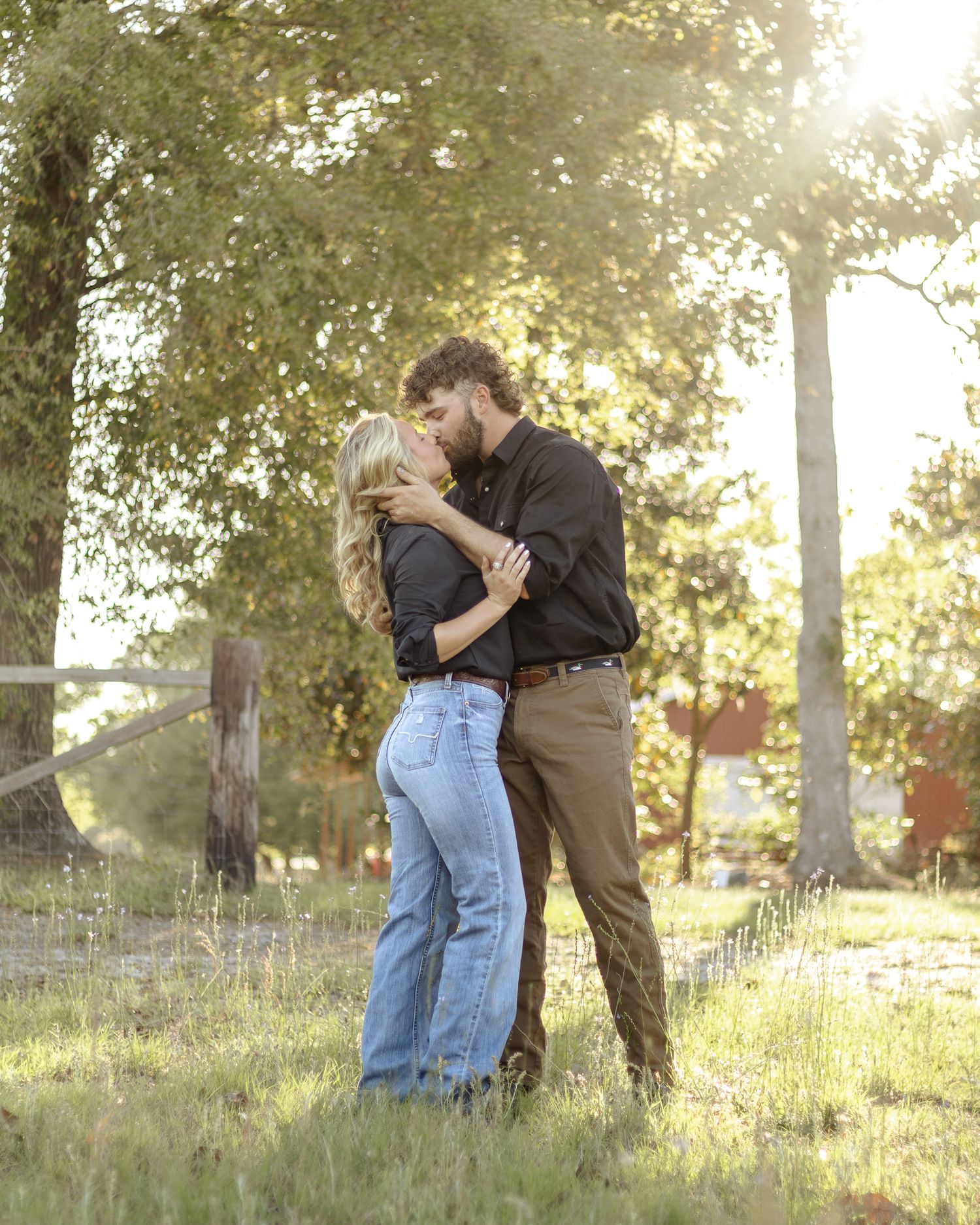 A couple in jeans and button-up shirts shares a kiss in a sunlit grassy field beside a wooden fence, surrounded by tall trees.