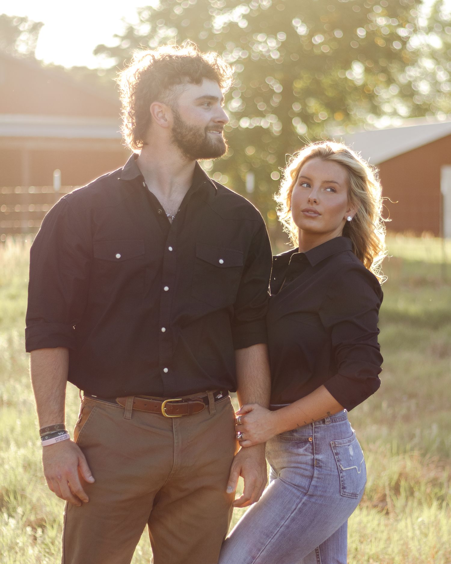 A bearded man and a blonde woman in matching dark button-up shirts hold hands in a sunlit grassy field with barns in the background.