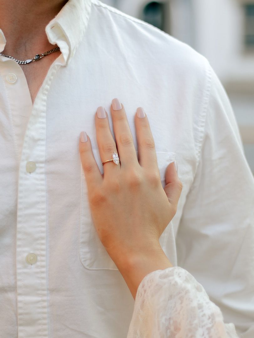 Woman's hand with a diamond engagement ring resting on the chest of a man in a white button-up shirt and silver chain necklace.