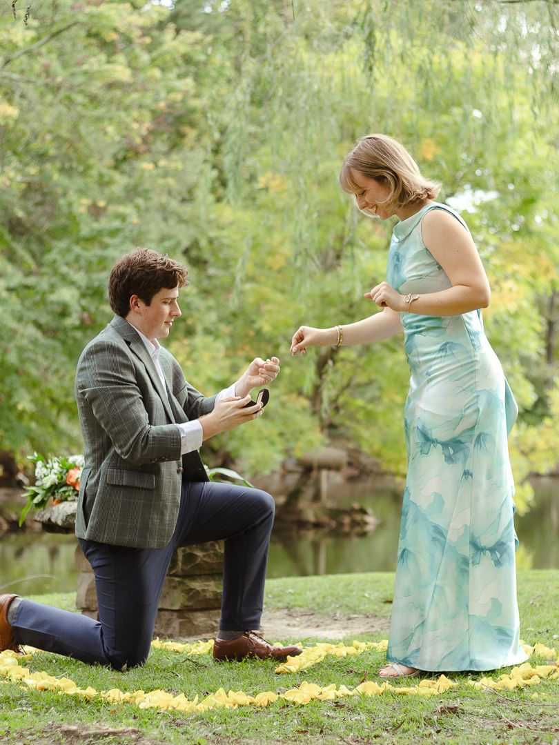 A man in a gray blazer kneels on a ring of yellow petals to propose to a woman in a blue dress beside a pond.