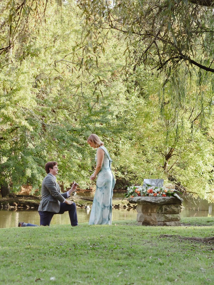 Man kneeling on one knee proposing to a woman in a long blue dress on a grassy lawn near a pond, with a floral-topped stone pedestal nearby.