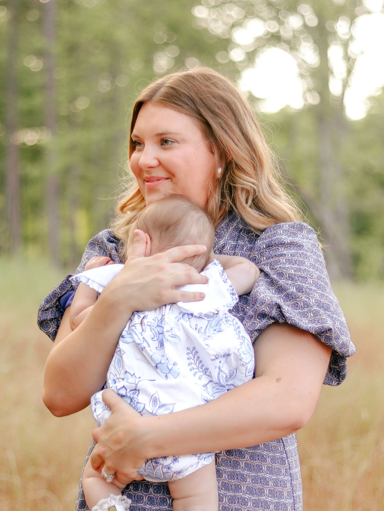 Smiling woman in a purple patterned dress cradles a baby in a blue floral outfit while standing in a sunlit grassy field with tall trees.