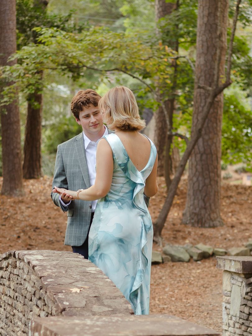 Man in a gray blazer holds hands with a woman in a flowing blue dress beside a stone bridge in a sunlit pine forest.