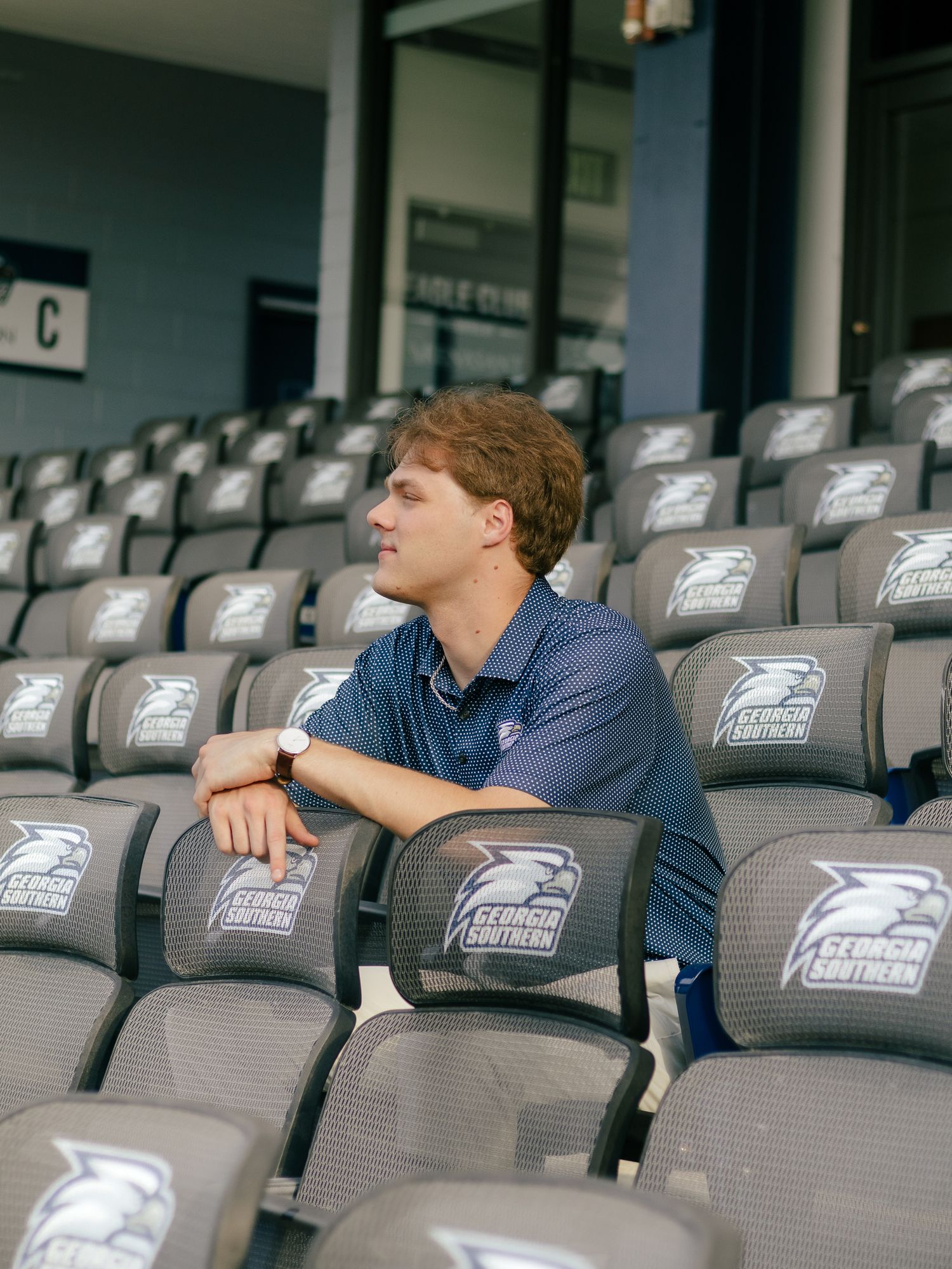 Young man in a navy polo sits on gray Georgia Southern stadium seats with eagle logos, resting his arm on the chair back.
