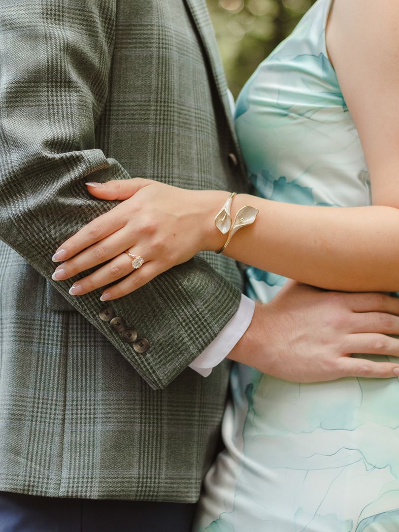 Close-up of a couple embracing, her hand with a diamond engagement ring and gold leaf bracelet resting on his green plaid blazer.