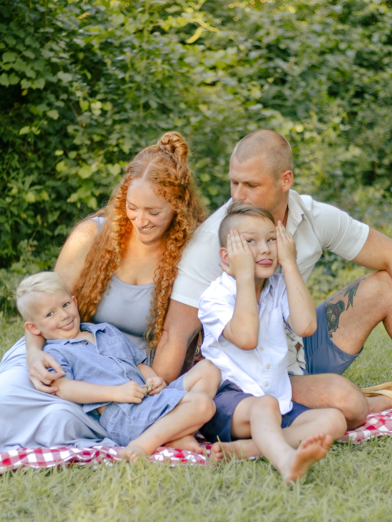 Smiling family of four with two young boys sitting together on a red checkered picnic blanket on green grass in a sunny park.